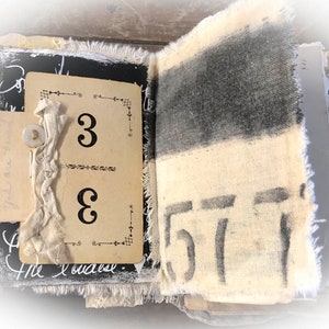 May include: An open journal with a black and white fabric cover. The pages are decorated with fabric scraps, a button, and a card with the number "3" written on it. The journal is open to a page with the number "57" written on it.