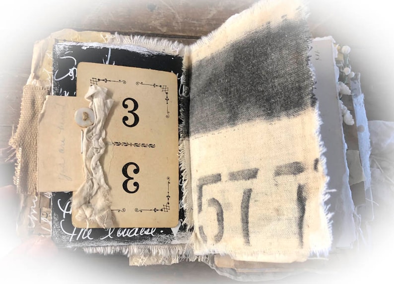 May include: An open journal with a black and white fabric cover. The pages are decorated with fabric scraps, a button, and a card with the number "3" written on it. The journal is open to a page with the number "57" written on it.