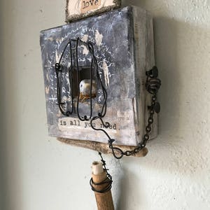 May include: A mixed media artwork featuring a bird in a cage, a scroll, and a crown with the word "love" on it. The piece is made from wood, metal, and paper. The background is a white wall.