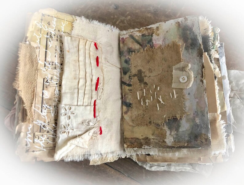 May include: An open journal with pages made of various fabrics, including burlap, lace, and linen. The pages are decorated with stitching, a button, and handwritten music notes.