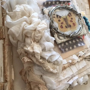 May include: A vintage-style journal with a white butterfly encased in wire and a white fabric cover. The journal is decorated with lace, fabric scraps, and a small button.
