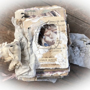 May include: A vintage journal with a distressed, layered look. The cover is made of paper and fabric, with a small, framed image of a cherub. The journal is tied with a pink ribbon and has a worn, rustic appearance.