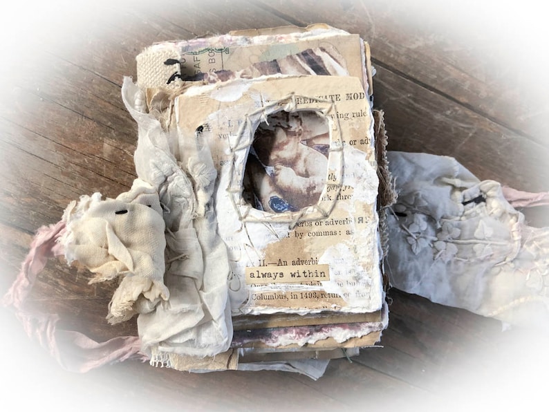 May include: A vintage journal with a distressed, layered look. The cover is made of paper and fabric, with a small, framed image of a cherub. The journal is tied with a pink ribbon and has a worn, rustic appearance.