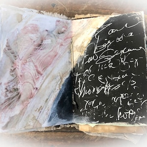 May include: An open journal with a black cover and white writing. The left page has a pink and white abstract design. The journal is on a wooden surface.