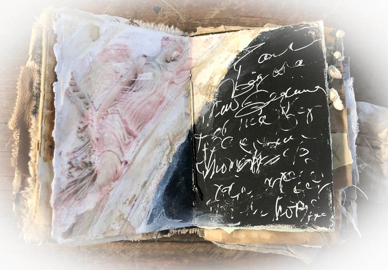 May include: An open journal with a black cover and white writing. The left page has a pink and white abstract design. The journal is on a wooden surface.