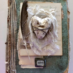 May include: A vintage book with a white heart-shaped rock wrapped in wire and fabric. The book has a blue cover and a striped fabric spine. The text "human beings" is visible on a page inside the book. A small metal tag with the word "spirit" is attached to the book.