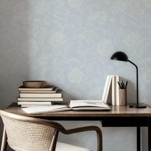 May include: A wooden desk with books, a lamp, and a wooden bowl. The desk is set against a light blue floral wallpaper. A black desk lamp is on the right side of the desk. A stack of books and a wooden bowl are on the left.