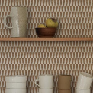 May include: A wallpaper featuring a repeating pattern of brown trapezoids on a cream background. The wallpaper is displayed behind two wooden shelves holding a ceramic pitcher, a wooden bowl with two yellow fruits, and stacks of ceramic mugs and bowls.