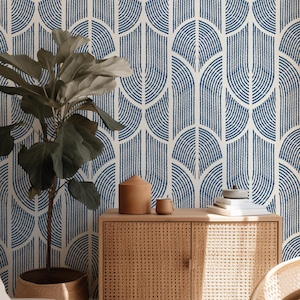 May include: A room interior featuring a blue and white patterned wallpaper with an Art Deco design. A wooden cabinet with a woven front holds a ceramic jar, cup, and books. A potted plant in a woven basket is in the foreground.