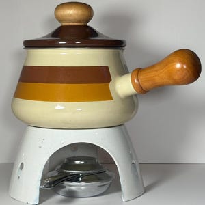 Può includere: Una pentola per fondue in smalto vintage con manico e coperchio in legno. La pentola ha una base bianca con strisce marroni e arancioni. La pentola è appoggiata su un supporto in metallo bianco con un bruciatore.