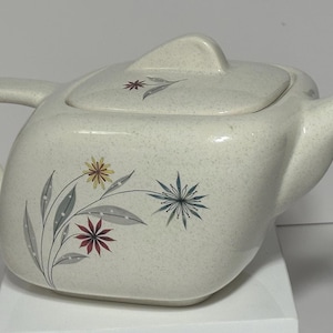 Może przedstawiać: Biały ceramiczny czajnik z kwadratowym korpusem i pokrywką. Czajnik ma kwiatowy wzór z szarymi, czerwonymi, żółtymi i niebieskimi kwiatami.