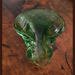 May include: A green glass bowl with a wide, shallow top and a flared base. The bowl has a bubbly texture and is made of transparent green glass. The bowl is likely a decorative piece, suitable for display or holding small items.