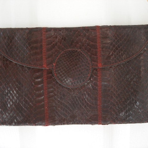 May include: A dark brown clutch with a snakeskin pattern, featuring a gold-colored frame and chain strap. Red stitching accents the design. The clutch has a flap closure with a circular detail.
