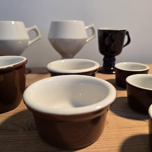 May include: A collection of ceramic kitchenware including several small brown and white ramekins, two white mugs with angular handles, and one dark brown pedestal mug. The items are arranged in a group, featuring smooth, glazed surfaces.
