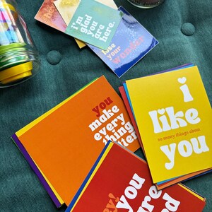 Peut inclure: Un assortiment de cartes de vœux colorées avec des messages tels que "I like you" et "You make everything better." Les cartes sont dans des tons d'orange, de jaune, de rouge et de bleu, avec du texte blanc. Certaines cartes ont un symbole de cœur. Les cartes sont sur une surface verte.