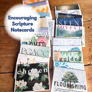 May include: A collection of encouraging scripture notecards with various designs and inspirational messages. The cards feature floral patterns, watercolor art, and phrases like "Chosen," "Peace," and "Flourishing." The cards are stacked on a wooden surface.