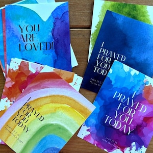 May include: A collection of watercolor greeting cards with inspirational messages. The cards feature vibrant splashes of color in blue, purple, orange, and green. Text includes "You Are Loved!" and "I Prayed For You Today."