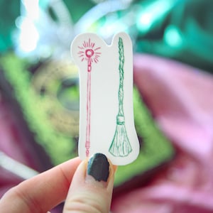 May include: A white sticker featuring two illustrations: a red wand with a starburst design and a green broom. The sticker is held against a blurred background of green and pink fabric.