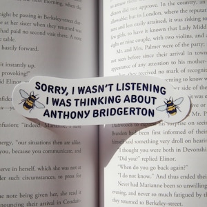 May include: White bookmark with the text "SORRY, I WASN'T LISTENING I WAS THINKING ABOUT ANTHONY BRIDGERTON" in black. Features bee illustrations on each end. Placed on an open book.