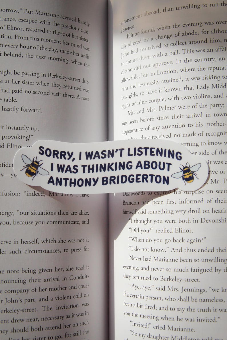 May include: White bookmark with the text "SORRY, I WASN'T LISTENING I WAS THINKING ABOUT ANTHONY BRIDGERTON" in black. Features bee illustrations on each end. Placed on an open book.