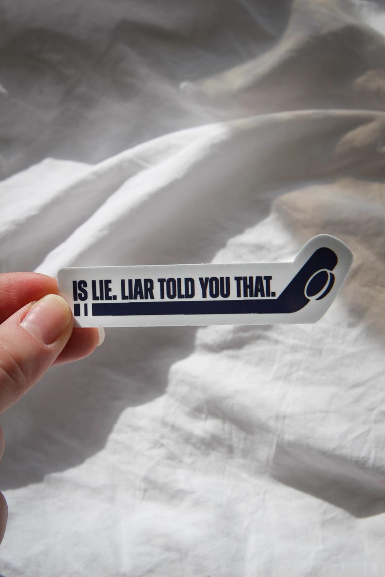 May include: A white sticker with the text "IS LIE. LIAR TOLD YOU THAT." in navy blue. The sticker has a stylized airplane tail design on the right side. The sticker is held against a white, wrinkled fabric background.