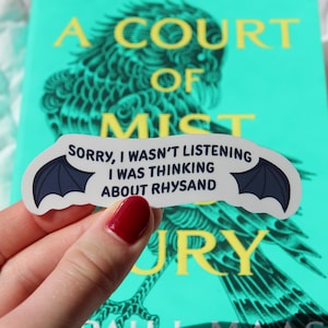 Puede incluir: Una pegatina con el texto "SORRY, I WASN'T LISTENING I WAS THINKING ABOUT RHYSAND" y un dise&ntilde;o de ala de murci&eacute;lago. La pegatina se sostiene frente a un libro titulado "A Court of Mist and Fury" de Sarah J. Maas.
