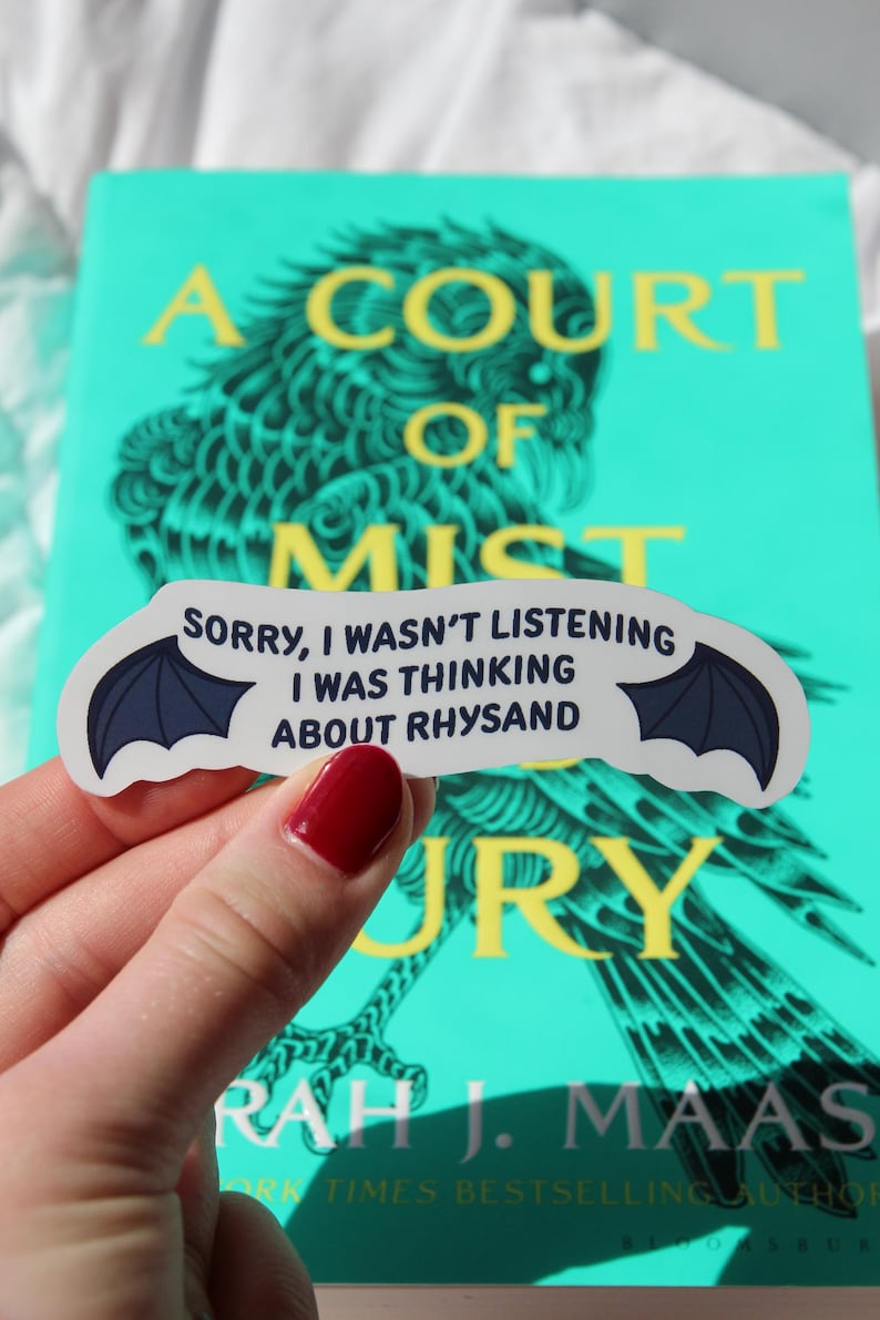 Puede incluir: Una pegatina con el texto "SORRY, I WASN'T LISTENING I WAS THINKING ABOUT RHYSAND" y un dise&ntilde;o de ala de murci&eacute;lago. La pegatina se sostiene frente a un libro titulado "A Court of Mist and Fury" de Sarah J. Maas.