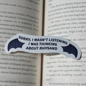 Puede incluir: Un marcap&aacute;ginas blanco con un dise&ntilde;o de alas de murci&eacute;lago azul oscuro y el texto "SORRY, I WASN'T LISTENING I WAS THINKING ABOUT RHYSAND" en negro. El marcap&aacute;ginas est&aacute; colocado en las p&aacute;ginas de un libro.