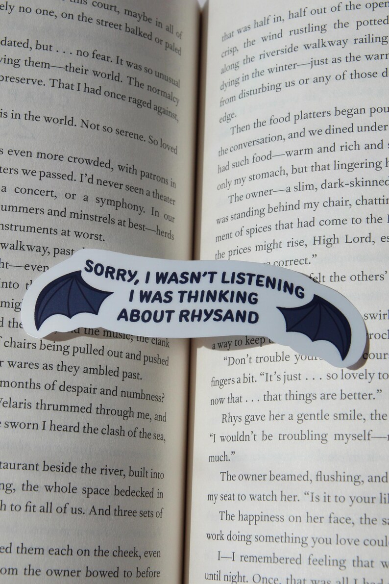 Puede incluir: Un marcap&aacute;ginas blanco con un dise&ntilde;o de alas de murci&eacute;lago azul oscuro y el texto "SORRY, I WASN'T LISTENING I WAS THINKING ABOUT RHYSAND" en negro. El marcap&aacute;ginas est&aacute; colocado en las p&aacute;ginas de un libro.