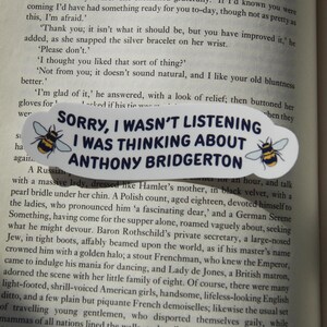 May include: A white, oval sticker with the text "SORRY, I WASN'T LISTENING I WAS THINKING ABOUT ANTHONY BRIDGERTON" in black. The sticker has a cartoon bee on each side. The sticker is on a book page.
