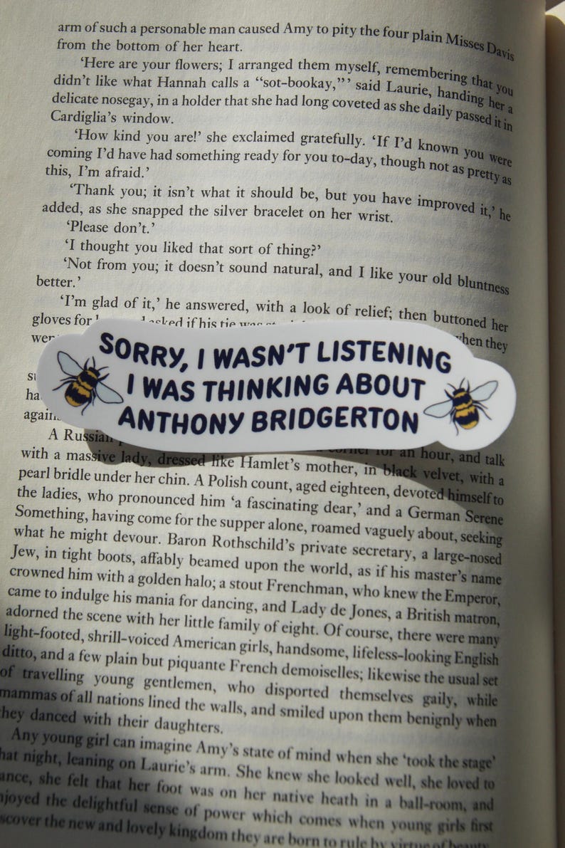 May include: A white, oval sticker with the text "SORRY, I WASN'T LISTENING I WAS THINKING ABOUT ANTHONY BRIDGERTON" in black. The sticker has a cartoon bee on each side. The sticker is on a book page.