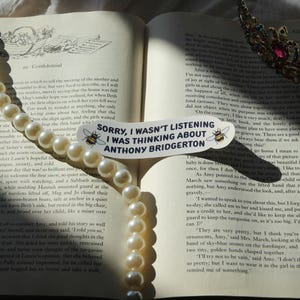 May include: An open book with a pearl necklace draped across the pages. A sticker with the text "Sorry, I wasn't listening I was thinking about Anthony Bridgerton" is on the page. A jeweled crown rests on the right page.