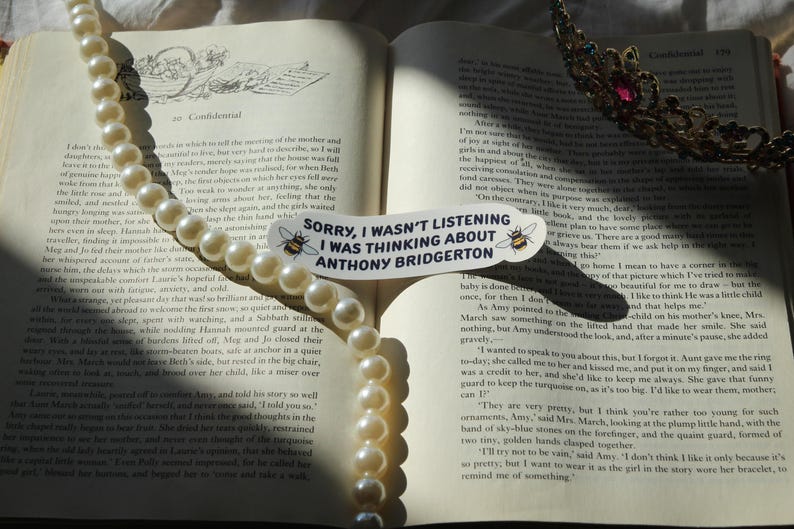 May include: An open book with a pearl necklace draped across the pages. A sticker with the text "Sorry, I wasn't listening I was thinking about Anthony Bridgerton" is on the page. A jeweled crown rests on the right page.