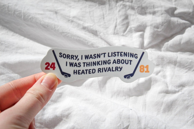 May include: A white sticker with the text "SORRY, I WASN'T LISTENING I WAS THINKING ABOUT HEATED RIVALRY." The sticker has two hockey sticks and the numbers 24 and 81 in red and orange. The sticker is held by a hand.