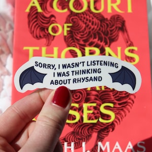 Puede incluir: Una pegatina con alas de murci&eacute;lago y el texto "Sorry, I wasn't listening, I was thinking about Rhysand" se sostiene frente a un libro titulado "A Court of Thorns and Roses" de H.J. Maas. La portada del libro es roja con texto amarillo.