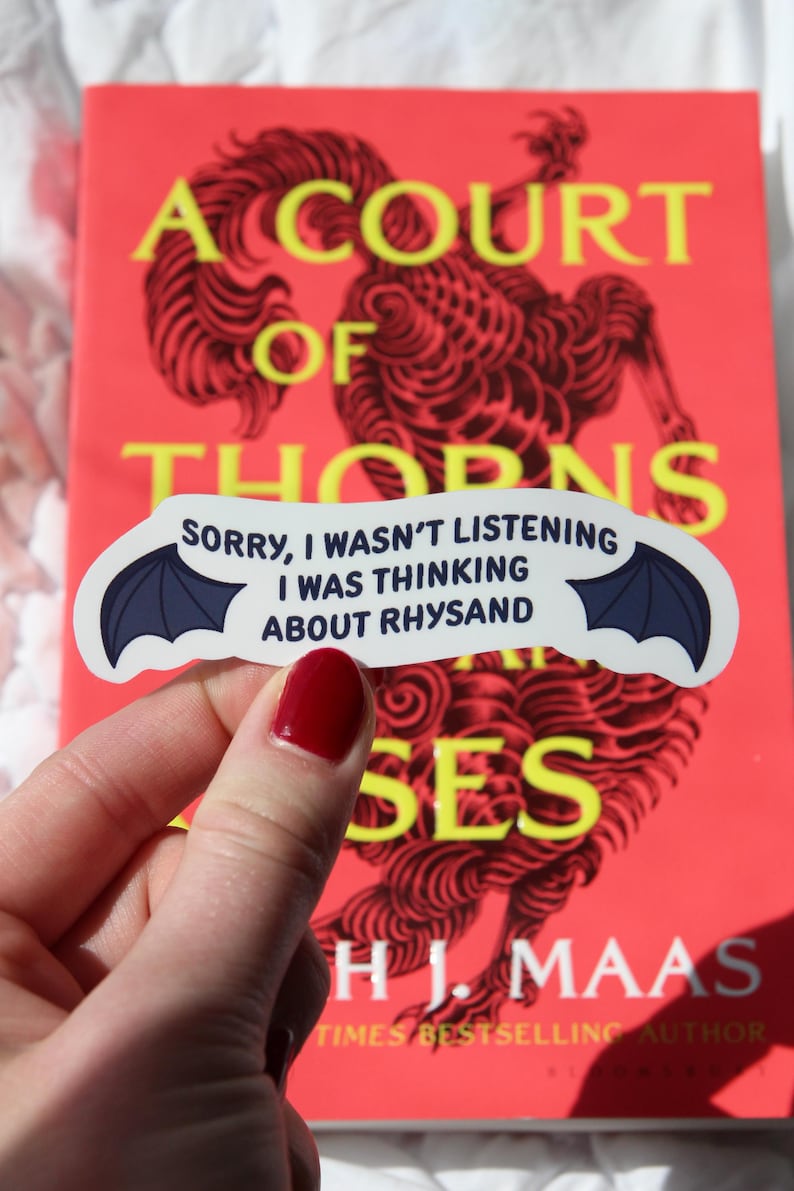 Puede incluir: Una pegatina con alas de murci&eacute;lago y el texto "Sorry, I wasn't listening, I was thinking about Rhysand" se sostiene frente a un libro titulado "A Court of Thorns and Roses" de H.J. Maas. La portada del libro es roja con texto amarillo.