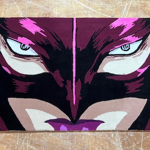 May include: A close-up of a burgundy and black rug with a stylised image of a face. The face has pink accents and large, white eyes.