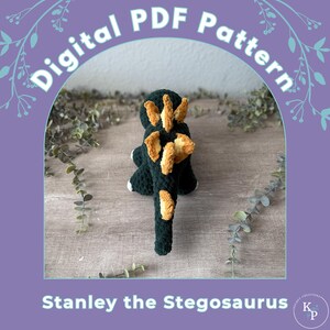 Op de afbeelding: Een handgemaakt, donkergroen gehaakt stegosaurus speelgoed met bruine platen op de rug en staart. Het speelgoed heeft witte accenten op de voeten. De afbeelding bevat de tekst "Digital PDF Pattern" en "Stanley the Stegosaurus".
