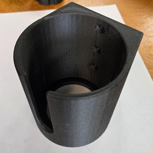 May include: A black, 3D-printed cup holder with a unique, angular design. The holder features a cylindrical interior and a square top, with a cutout on one side. Three small holes are visible on the interior wall.