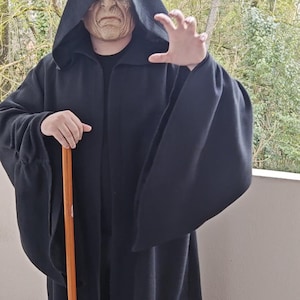 Peut inclure: Une longue robe noire à capuche avec de larges manches, portée avec un masque. Le masque est de couleur pâle avec des traits faciaux détaillés. Un bâton en bois est tenu dans une main. Le costume est probablement pour le cosplay ou Halloween.