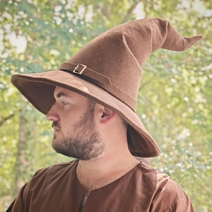 Pode incluir: Um chapéu de mago castanho com aba larga e ponta. O chapéu tem uma correia com uma fivela prateada. O chapéu é feito de um material semelhante a feltro. O chapéu é usado por uma pessoa.