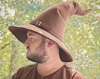 May include: A brown wizard hat with a wide brim and a pointed tip. The hat has a strap with a silver buckle. The hat is made of a felt-like material. The hat is worn by a person.