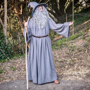 Pode incluir: Uma pessoa com uma fantasia de mago cinzento, completa com uma túnica comprida, chapéu e bengala. A fantasia inclui uma longa barba cinzenta e um cinto castanho. O mago está em pé num caminho pavimentado numa área arborizada.