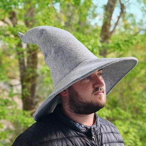 Puede incluir: Un sombrero de mago de fieltro gris con un ala ancha y una corona alta y puntiaguda. El sombrero lo lleva puesto una persona con barba.