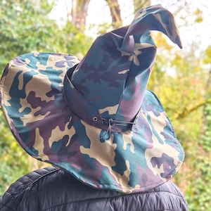 May include: A camouflage-patterned witch hat with a wide brim and a pointed top. The hat features a mix of green, blue, purple, and tan colors. A dark strap encircles the base of the hat, and a black jacket is visible beneath.