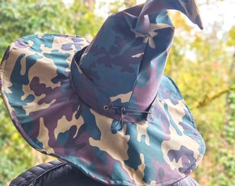 May include: A camouflage-patterned witch hat with a wide brim and a pointed top. The hat features a mix of green, blue, purple, and tan colors. A dark strap encircles the base of the hat, and a black jacket is visible beneath.