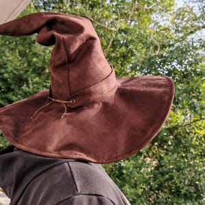 Pode incluir: Um chapéu pontiagudo castanho com uma aba larga e flexível e uma copa alta ligeiramente caída. O chapéu apresenta uma faixa decorativa em torno da base da copa, fixada com um cordão de couro fino através de ilhós metálicos na frente.