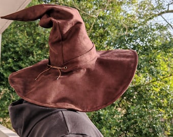 May include: A brown pointed hat with a wide, flexible brim and a tall, slightly slumped crown. The hat features a decorative band around the base of the crown, secured with a thin leather cord threaded through metal eyelets at the front.