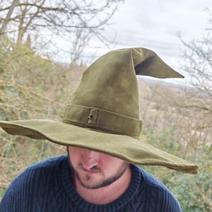 May include: A wide-brimmed, green felt wizard hat with a pointed crown and a brown leather buckle. The hat is being worn by a person with a beard.