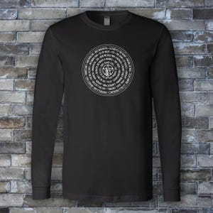 May include: Black long-sleeve shirt with a white circular graphic on the front. The graphic features a central symbol surrounded by text. The shirt is made of a soft material and has a crew neck.